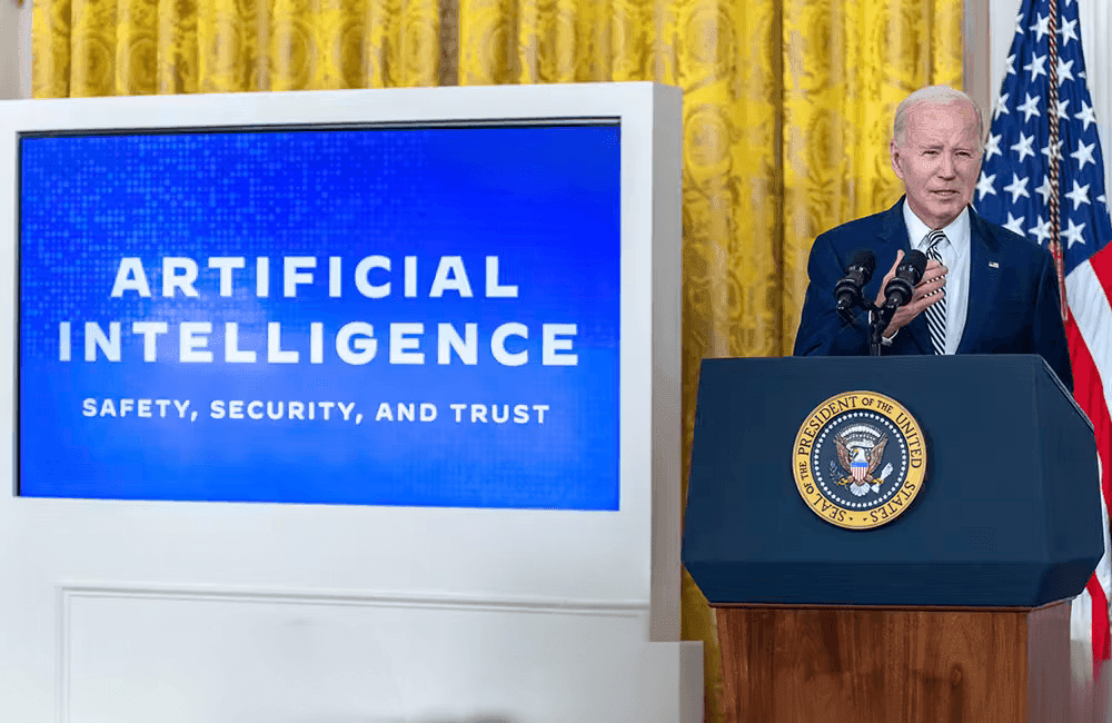 President Biden speaks at a podium about AI safety, security, and trust, a large screen displays the same message behind him.