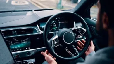 Person's hands on a Jaguar's self-driving steering wheel, digital dashboard displays speed and media options.