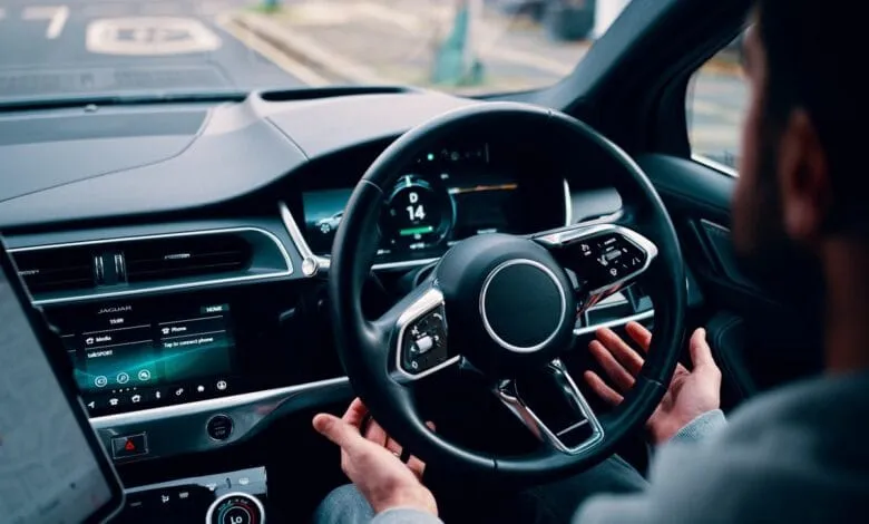 Person's hands on a Jaguar's self-driving steering wheel, digital dashboard displays speed and media options.