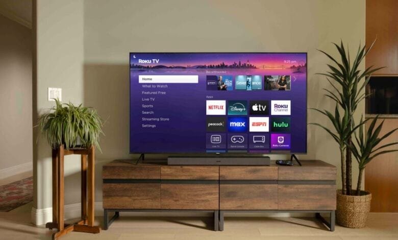 Roku TV home screen showing Netflix, Disney+, Apple TV, and other apps. The TV sits on a wooden media console in a modern living room.