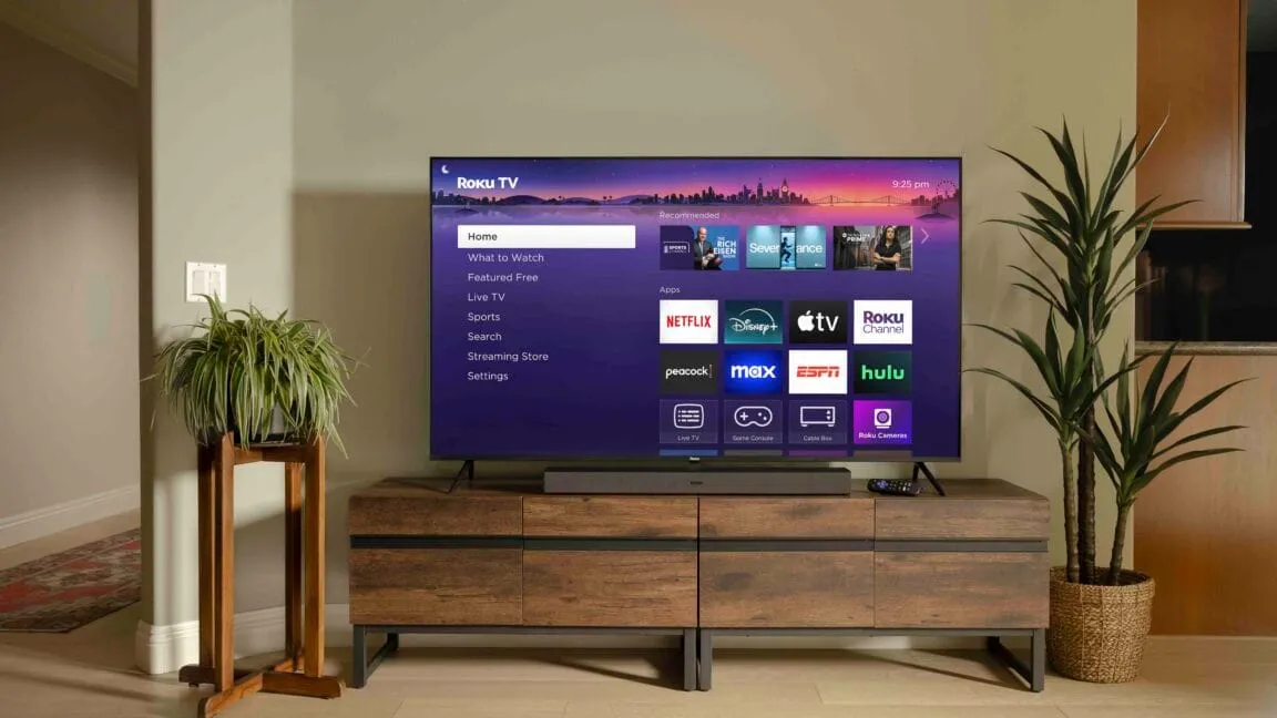 Roku TV home screen showing Netflix, Disney+, Apple TV, and other apps. The TV sits on a wooden media console in a modern living room.
