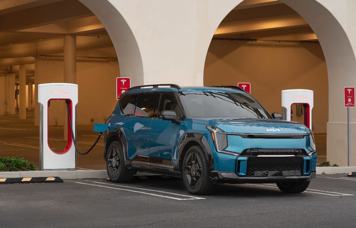 A teal Kia EV6 SUV charges at a Tesla Supercharger station in a modern parking garage.