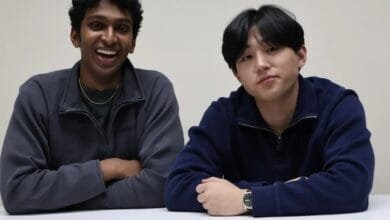 Two young men, one with dark curly hair and a joyful expression, the other with dark straight hair, sit side-by-side at a table.