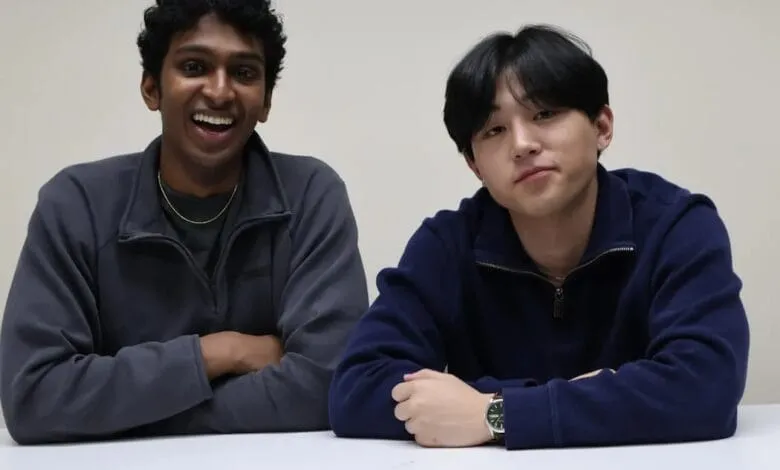 Two young men, one with dark curly hair and a joyful expression, the other with dark straight hair, sit side-by-side at a table.