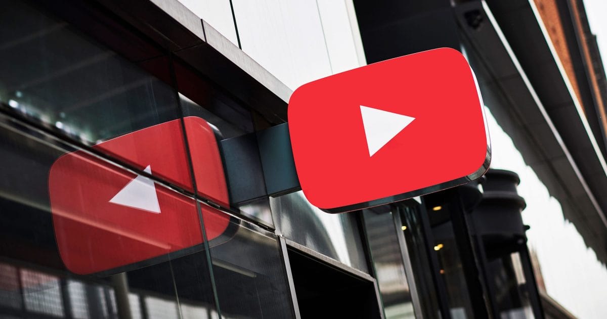 A bright red YouTube logo with a white play button is mounted on a modern building's glass exterior. Its reflection is visible.