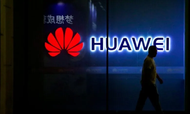 Brightly lit Huawei logo in red and white on a dark building at night. A person walks past.