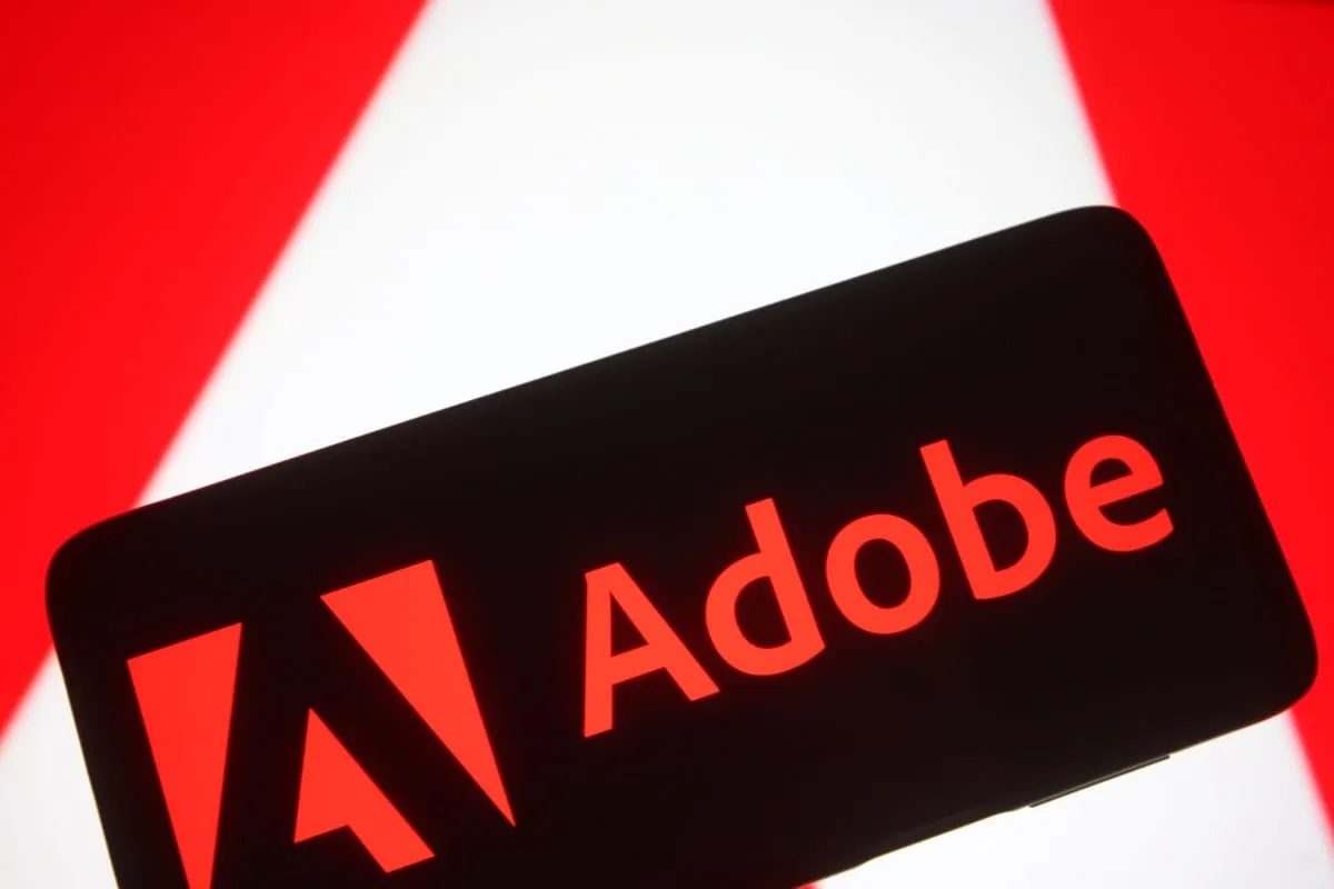 Close-up of a smartphone displaying the Adobe logo in bright red against a black screen. Blurred red and white geometric shapes are in the background.