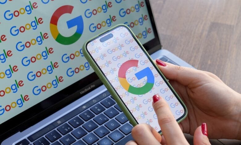 Close-up of hands using a smartphone displaying the Google logo. The phone rests on a laptop showing a Google-logo background.