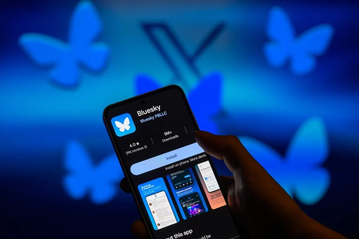 Close-up of a smartphone displaying the Bluesky social media app download page. Blue butterflies are blurred in the background.