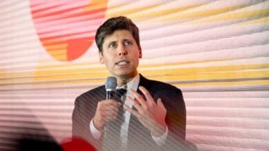 Sam Altman speaks into a microphone at a conference. The background is a blurred, colorful screen.