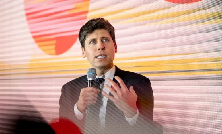Sam Altman speaks into a microphone at a conference. The background is a blurred, colorful screen.