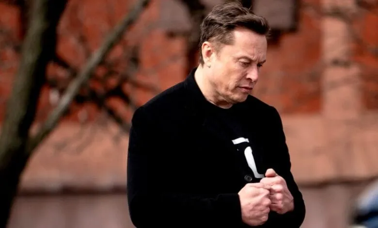 Elon Musk, CEO of Tesla and SpaceX, stands outdoors with a serious expression, wearing a black jacket and shirt.