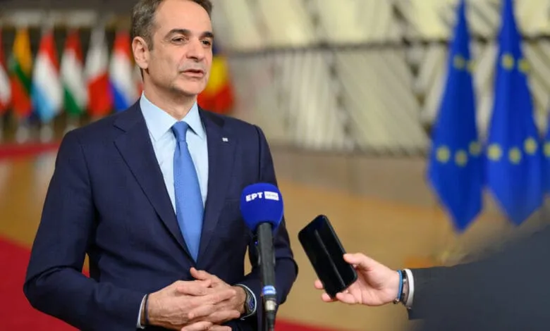 Kyriakos Mitsotakis, Prime Minister of Greece, speaks to reporters, holding his hands together. Microphones and a phone are visible.