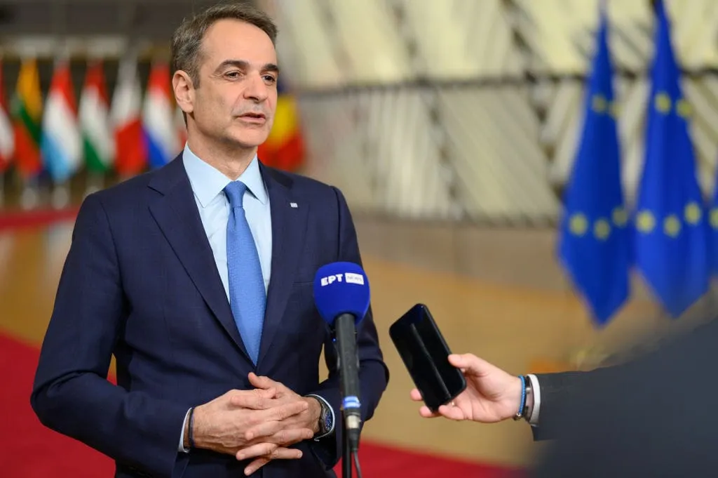 Kyriakos Mitsotakis, Prime Minister of Greece, speaks to reporters, holding his hands together. Microphones and a phone are visible.