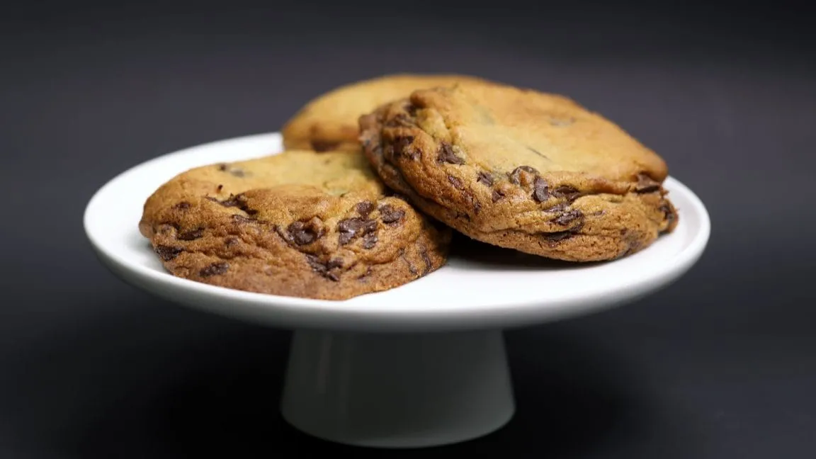Three large, golden-brown chocolate chip cookies sit on a small white pedestal cake stand against a dark background.