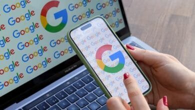Close-up of hands holding a smartphone displaying the Google logo. A laptop in the background also shows the Google logo.