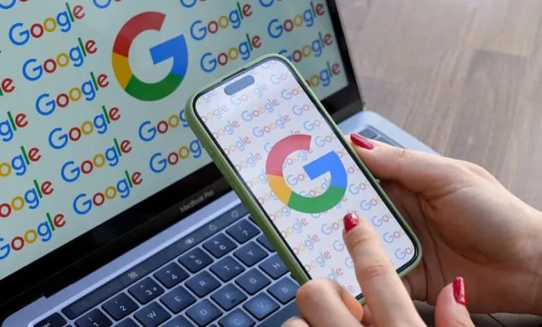 Close-up of hands holding a smartphone displaying the Google logo. A laptop in the background also shows the Google logo.