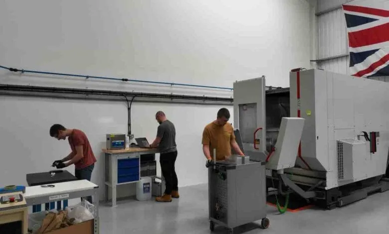 Three engineers work in a modern factory, using laptops and machinery. A British flag hangs in the background.