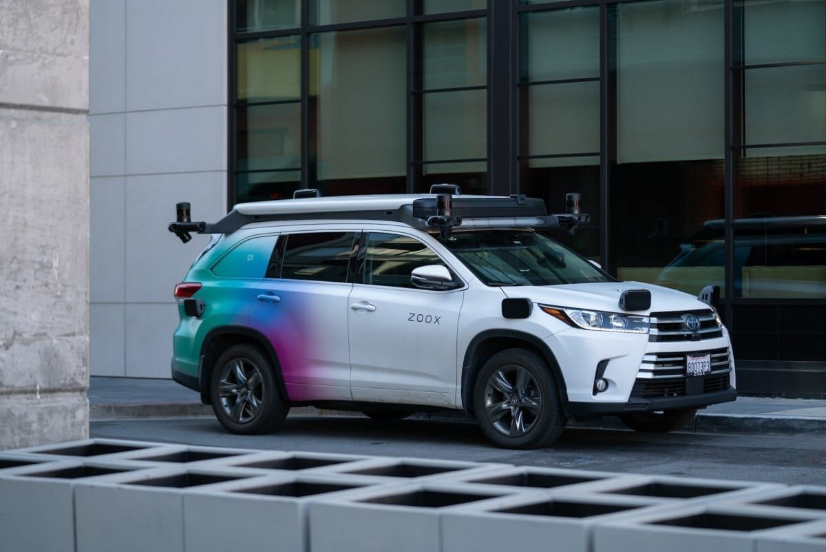 A white Zoox self-driving SUV is parked in front of a modern building. The car has various sensors on its roof.
