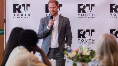 Prince Harry speaks at a Youth Power Fund summit, holding a microphone. He's wearing a gray suit and white shirt.