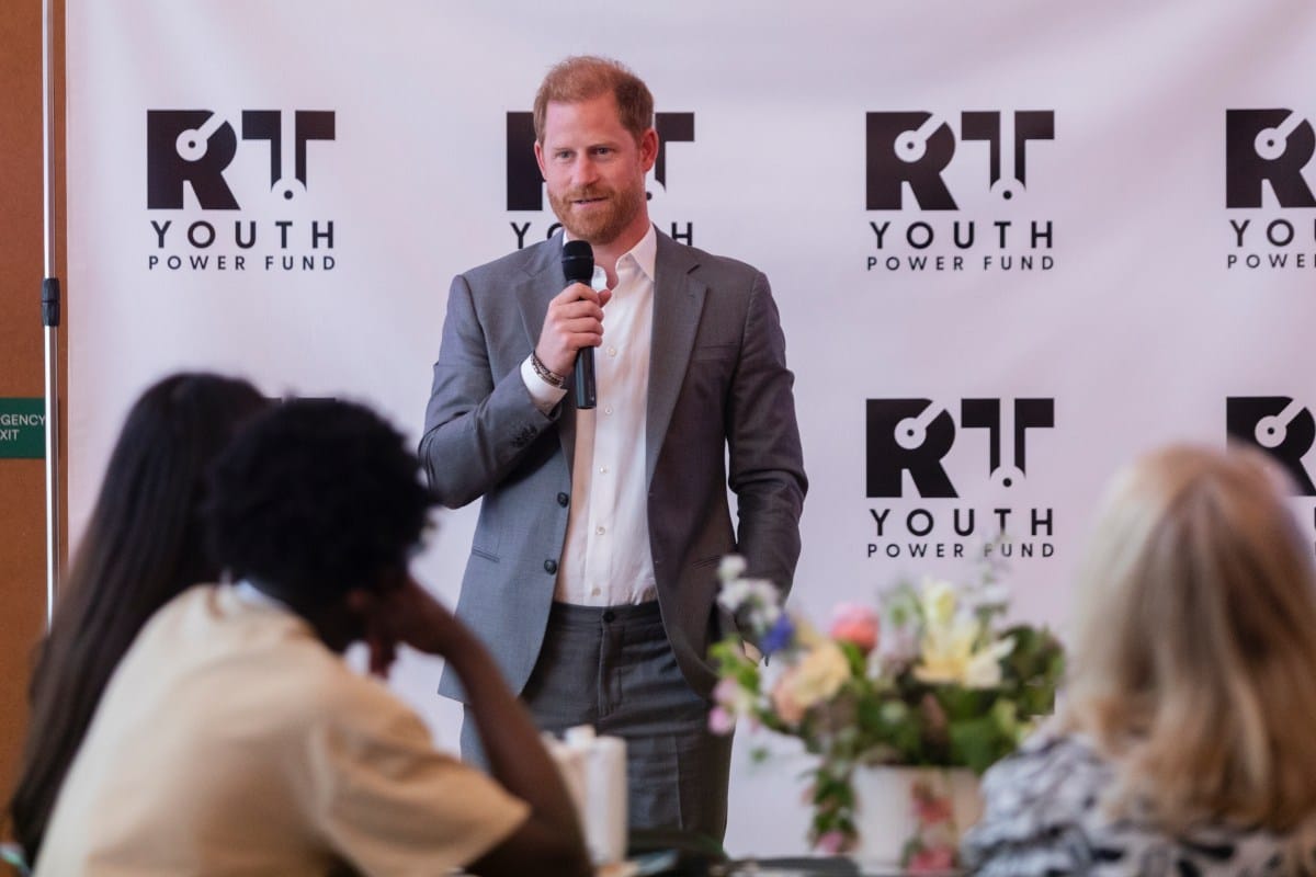 Prince Harry speaks at a Youth Power Fund summit, holding a microphone. He's wearing a gray suit and white shirt.
