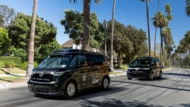 Two self-driving Volkswagen vans, branded with Uber, drive down a palm tree-lined street.