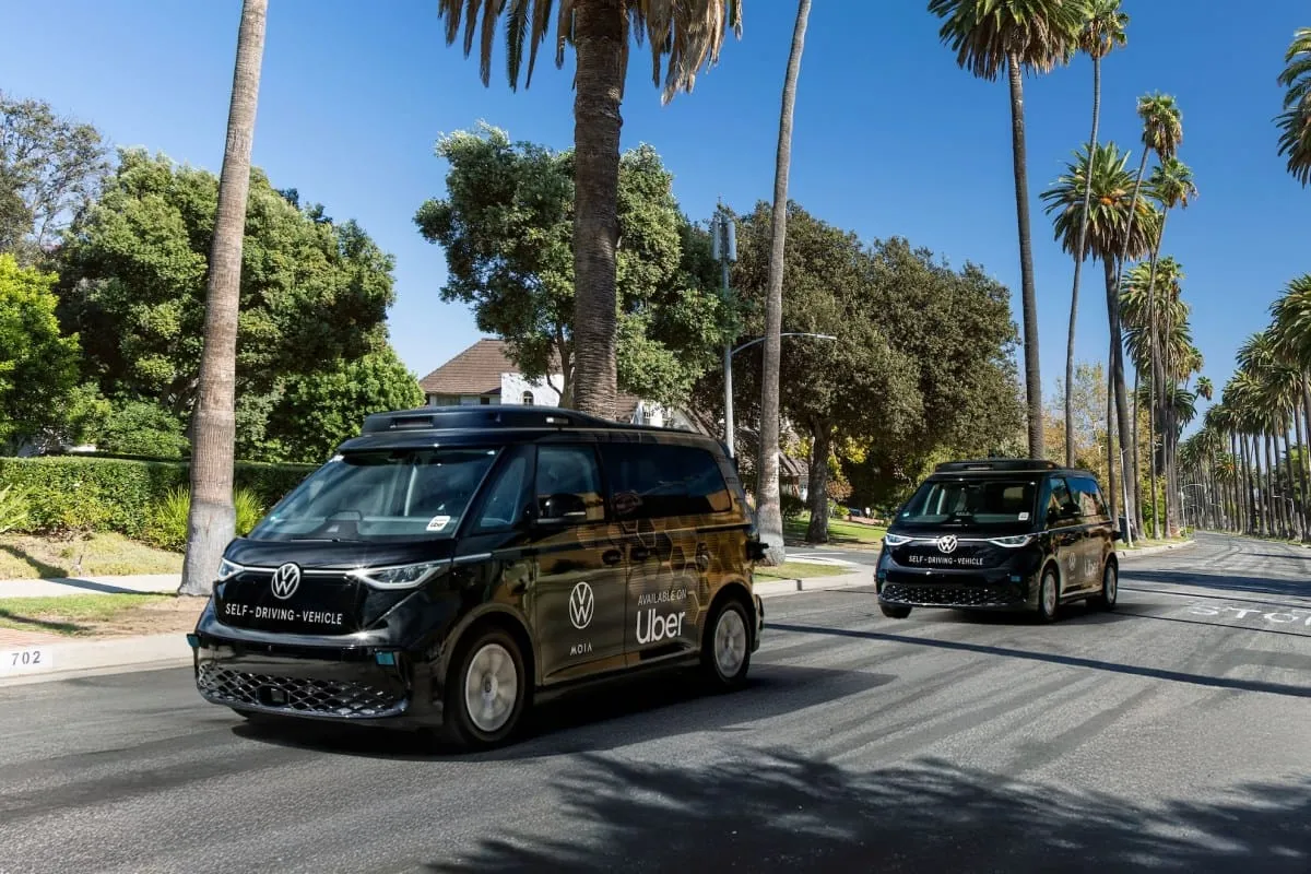 Two self-driving Volkswagen vans, branded with Uber, drive down a palm tree-lined street.