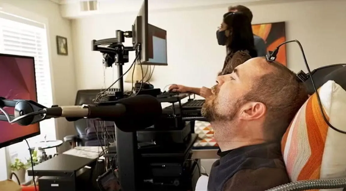 A man reclines in a chair with a device attached to his head, connected to computer equipment; two other people are in the background.