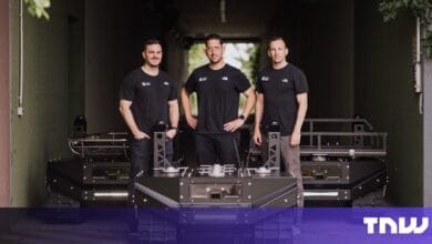 Three men stand proudly beside a large, dark-grey tracked robot in a garage setting.