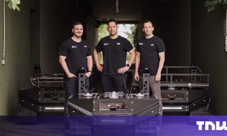 Three men stand proudly beside a large, dark-grey tracked robot in a garage setting.