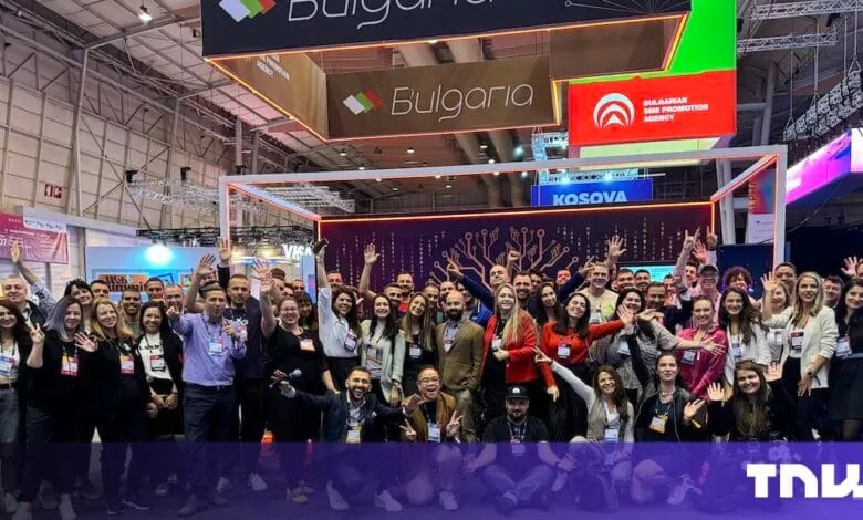 Large group of people at a tech conference, smiling and raising their hands, in front of various company booths.