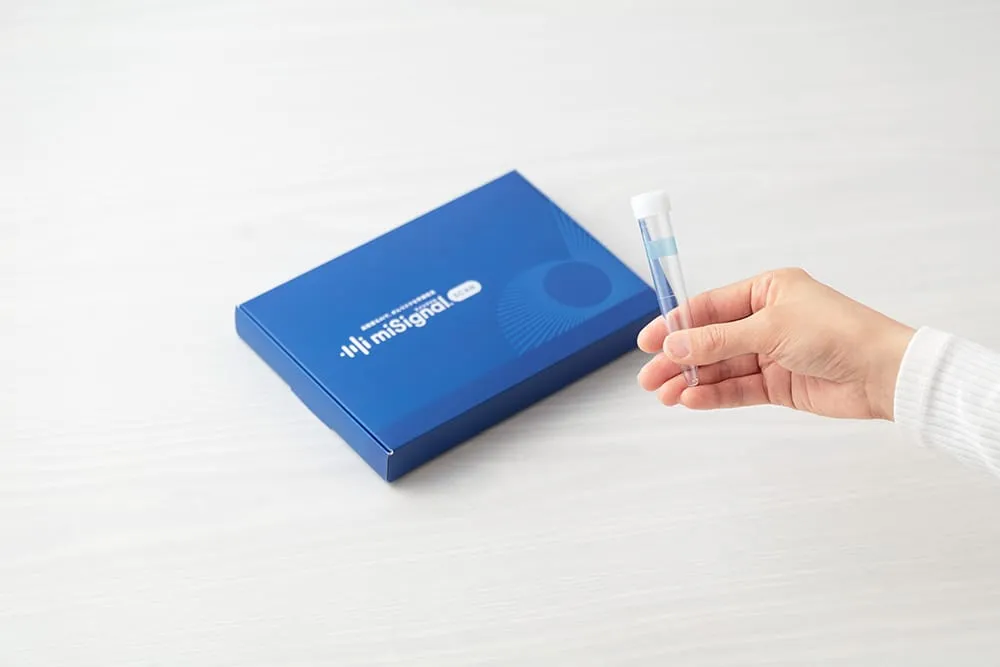 A hand holds a clear test tube next to a blue miSignal box. Medical testing kit.