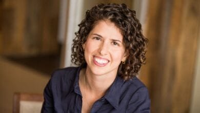Close-up portrait of Sarah Tavel, a smiling woman with curly brown hair, wearing a dark blue shirt.