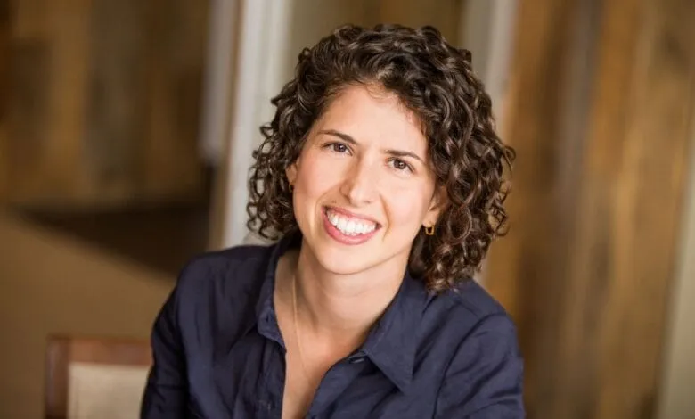 Close-up portrait of Sarah Tavel, a smiling woman with curly brown hair, wearing a dark blue shirt.
