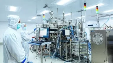 Two scientists in sterile suits monitor complex semiconductor manufacturing equipment in a pristine lab.