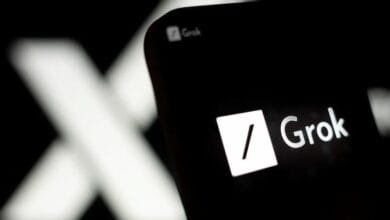 Close-up of a smartphone displaying the Grok AI app logo. A blurred 'X' is visible in the background.