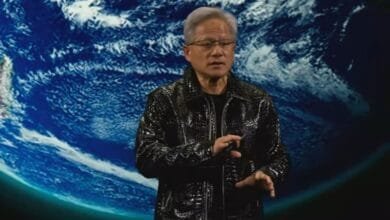A man in a black leather jacket gestures while standing in front of a backdrop showing Earth from space.