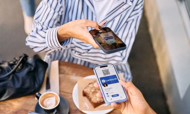 Two people scan QR codes on their smartphones at a cafe table. One phone displays a digital business card.