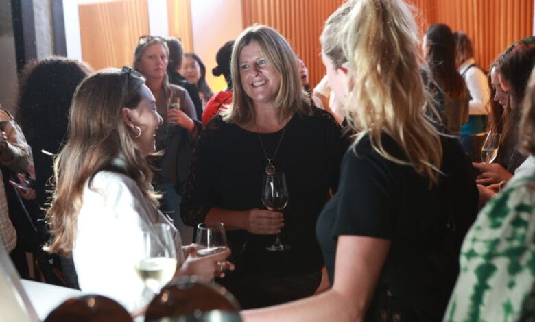Women at a social gathering, holding wine glasses and chatting. Warm lighting and a sophisticated atmosphere.