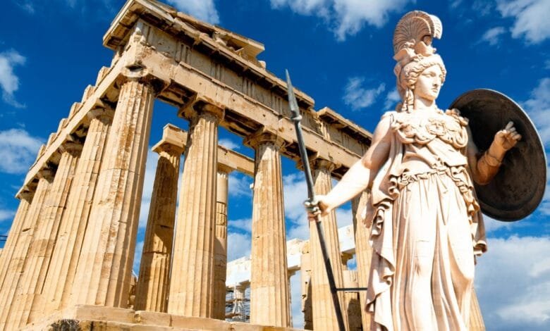 A statue of Athena, goddess of wisdom and war, stands before the ancient ruins of a Greek temple under a bright blue sky.