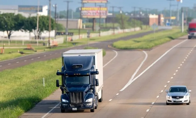 A dark-blue semi-truck and a silver sedan travel down a multi-lane highway, with billboards visible in the distance.