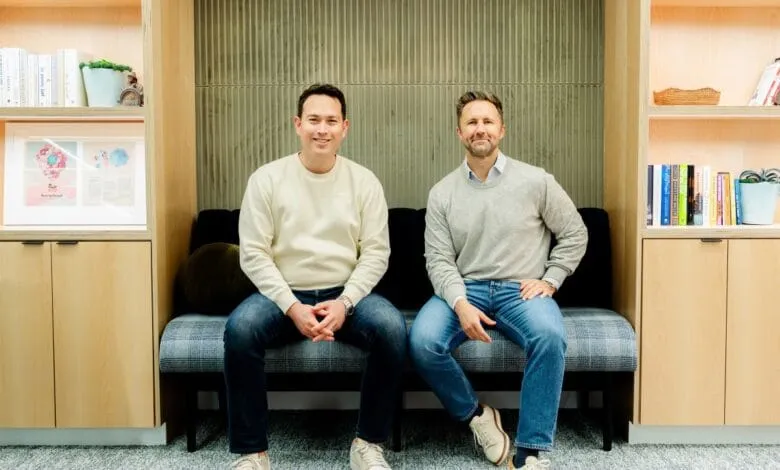 Two men smiling, sitting on a couch in a modern office. They are casually dressed and appear relaxed.
