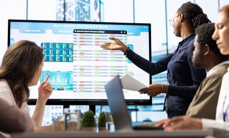 A woman leads a business meeting, presenting data on a large screen to colleagues. Charts and graphs display financial information.