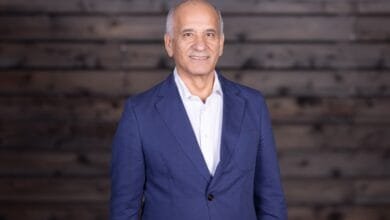 Smiling mature man, Faraj Aalaei, in a blue blazer stands against a dark wood background.