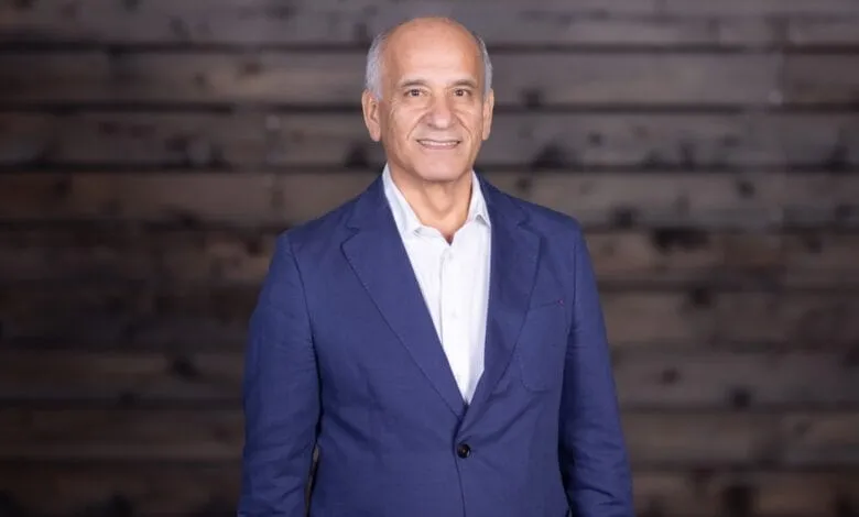 Smiling mature man, Faraj Aalaei, in a blue blazer stands against a dark wood background.