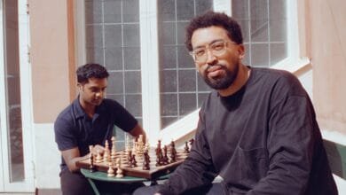 Two men play chess outdoors on a sunny day. One man wears glasses and a dark shirt; the other wears a dark polo shirt.