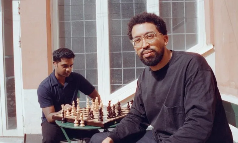 Two men play chess outdoors on a sunny day. One man wears glasses and a dark shirt; the other wears a dark polo shirt.
