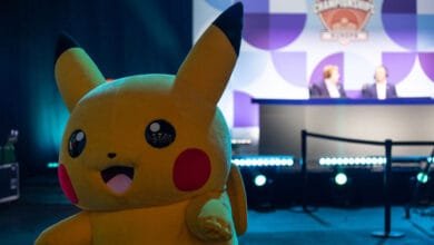 Pikachu mascot in front of a blurred background showing a Pokemon Championship stage with judges.