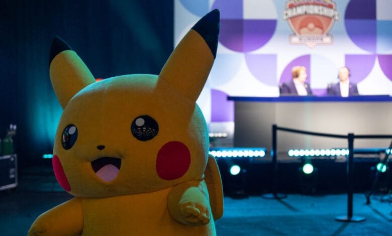 Pikachu mascot in front of a blurred background showing a Pokemon Championship stage with judges.