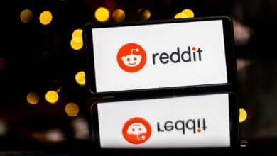 Smartphone displaying the Reddit logo: an orange alien mascot and 'reddit' in black text. Reflected on a dark surface.